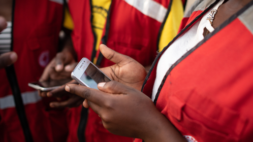 Three Ugandan Red Cross workers in Red Cross vests are looking at information on two mobile smartphones.