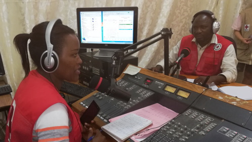 Two Red Cross workers - one male, one female - record a radio programme in a studio with a mic, computers and a sound deck.