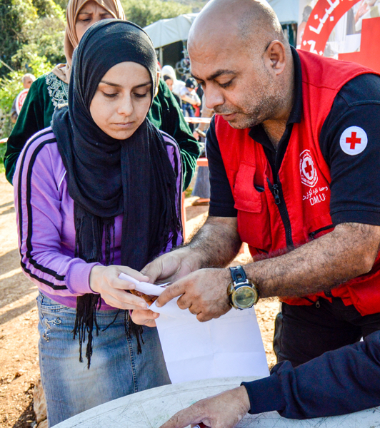 A male Red Cross worker helps a muslim woman with some paperwork at a camp. Another muslim woman waits in line behind her.