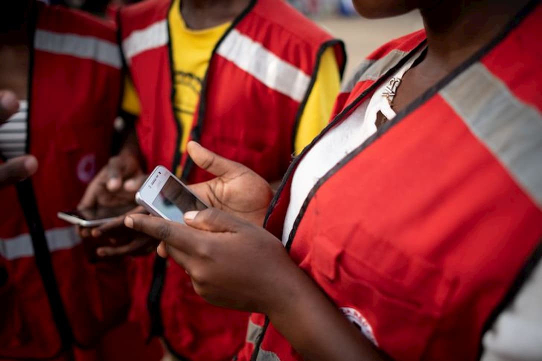 Three Ugandan Red Cross workers in Red Cross vests are looking at information on two mobile smartphones.