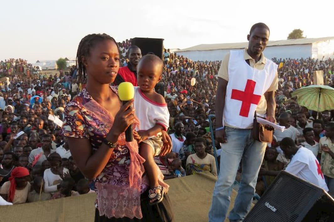 A woman holding a baby speaks into a microphone on stage whilst Red Cross workers and a large crowd watches behind her
